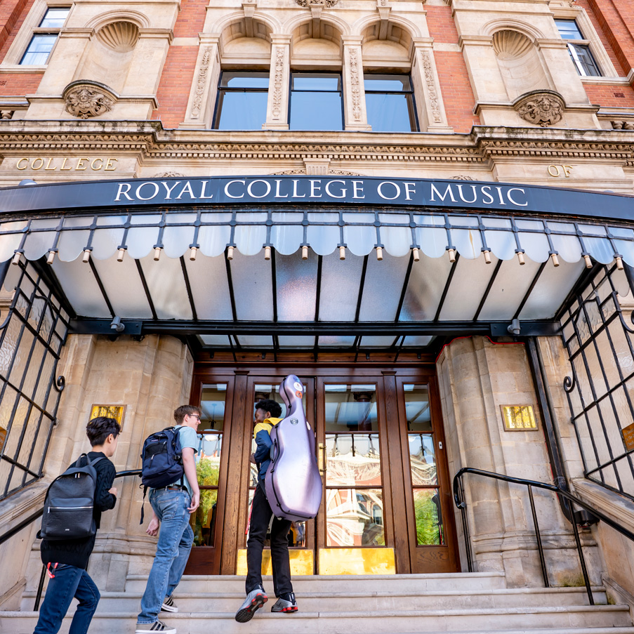 A group of students wearing casual clothes, with students holding instrument cases walking into the building of the Royal College of Music, an old, red brick, Victorian building, in the background.
